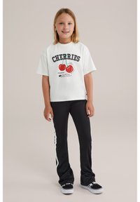 Balta medvilninė marškinė su "CHERRIES" grafika ir raudonų vyšnių dizainu, derinami su juodomis platėjančiomis kelnėmis, turinčiomis baltas šonines juosteles, ir juodais sportbačiais.