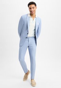 Helles blaues Anzug mit slim fit, strukturiertem Stoff, kombiniert mit einem hell gestreiften Poloshirt und beigen Slippern.