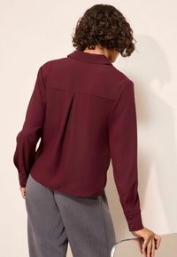 Bordowa bluzka z wywijanym kołnierzykiem, długimi rękawami i zapinanymi mankietami; posiada dodatkowy detal w postaci fałdy z tyłu. Połączona ze szarymi spodniami.