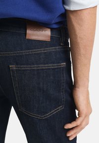 Person wearing dark blue denim jeans with brown leather GANT label on waistband and visible back pocket stitching, hand relaxed by side.