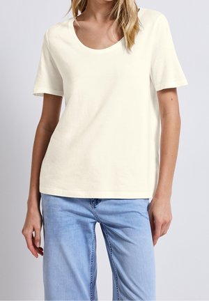 Kobieta w prostej białej bluzce z krótkim rękawem i okrągłym dekoltem, zestawiona z jasnoniebieskimi, wysokim stanem jeansami, na neutralnym tle.