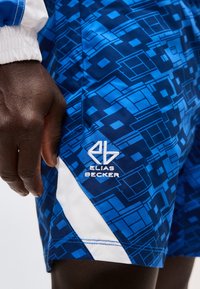 Χέρι χαλαρό δίπλα σε μπλε σορτς με γεωμετρικά λευκά σχέδια και κεντημένο λογότυπο "Elias Becker" στο αριστερό μηρό.