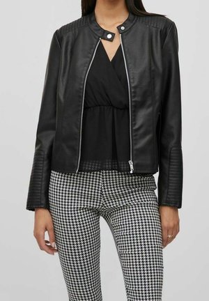 Frau trägt eine schwarze Lederjacke mit Reißverschluss, ein schwarzes V-förmiges Oberteil und schwarz-weiß karierte Hahnentritt-Hose.