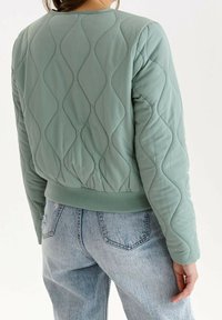 Person, der eine gequiltete, salbeigrüne Jacke und hellblaue Jeans trägt, von hinten gezeigt, vor einem schlichten weißen Hintergrund stehend.