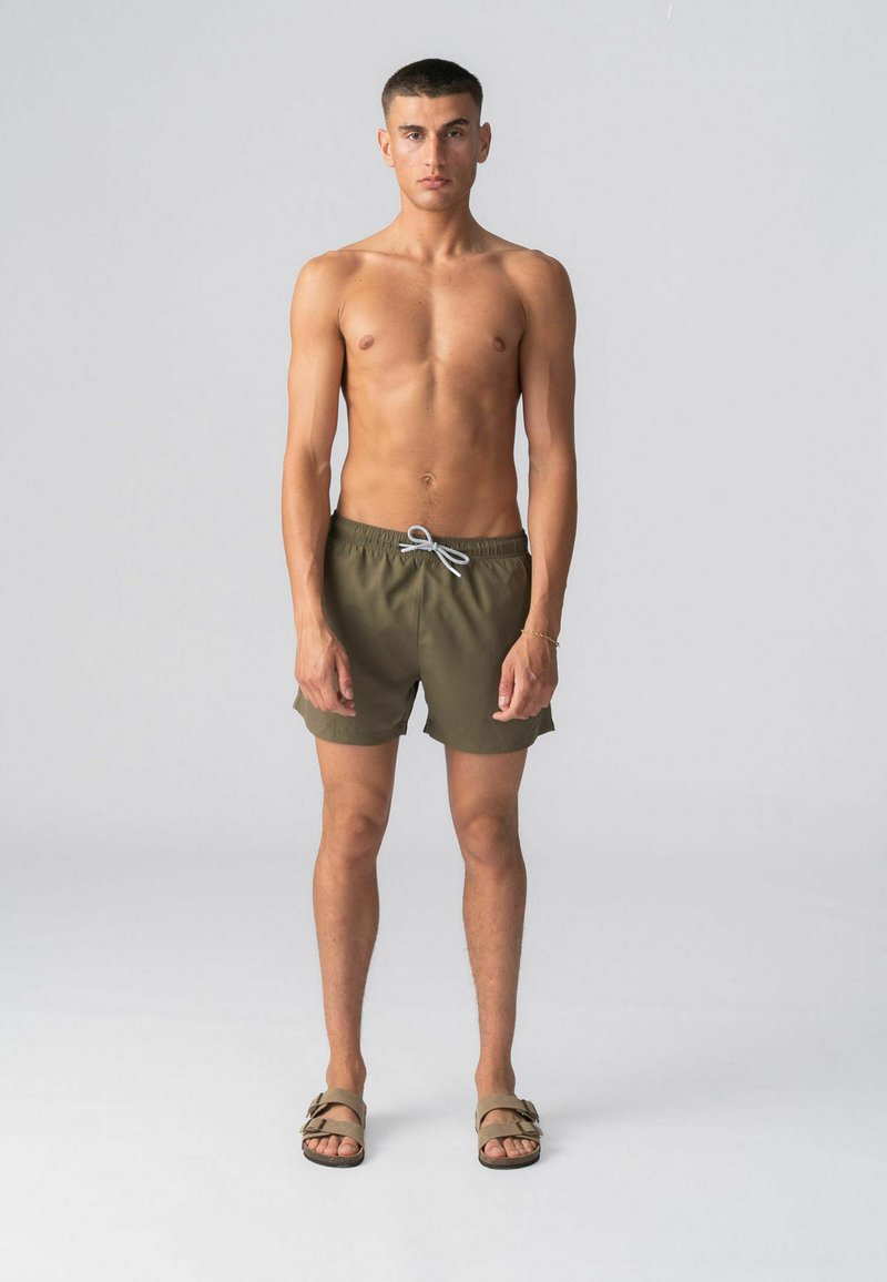 Jeune homme debout de face portant un short de bain vert olive avec un cordon de serrage et des sandales beiges à boucle, sur un fond gris uni.