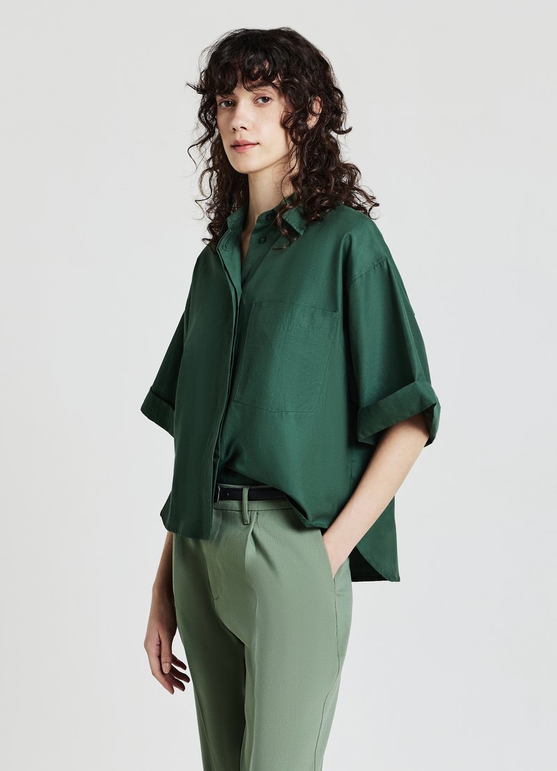 Oversized green button-up shirt with a smooth texture, short sleeves, and a chest pocket, paired with tailored light green pants.