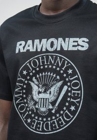 Melna kokvilnas T-kreklu ar nolietotu balto apdruku, kurā ir redzams apļveida emblēma ar ērgli, sikspārni un teksts "RAMONES" virsū.