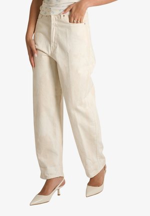 Donna che indossa jeans larghi a vita alta color crema, top bianco floreale e décolleté slingback beige con punta, mano in tasca.