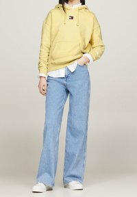 Felpa gialla con cappuccio, dotata di tasca frontale e logo, abbinata a jeans a gamba larga di colore blu chiaro. Le sneakers bianche completano l'outfit.