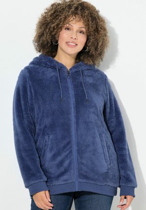 Kvinde med krøllet hår iført en blå fleece hoodie med lynlås og lommer, stående mod en ensfarvet hvid baggrund.