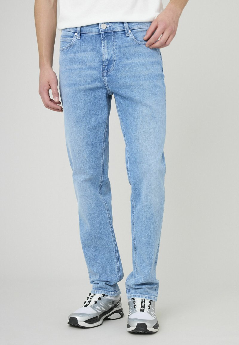 Jeans en denim bleu clair à coupe slim, présentant un design classique à cinq poches, des passants de ceinture et une fermeture à bouton. Portés avec des baskets grises.