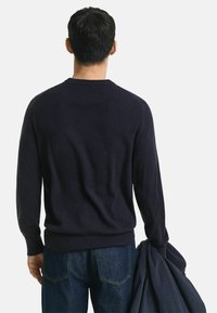 Marineblauer Strickpullover mit gerippten Bündchen und Saum, von hinten gezeigt, kombiniert mit Jeans und einem dunklen Oberbekleidungsstück, das über dem Arm hängt.