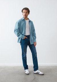 Camicia a righe blu e bianche con bottoni, t-shirt bianca, jeans blu e sneakers bianche con accenti grigi e neri, in piedi su uno sfondo neutro.