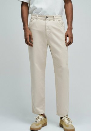 Hombre con pantalones beige de corte recto y zapatillas beige, de pie frente a un fondo gris claro y liso.