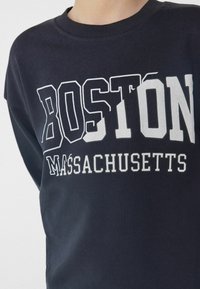 Tmavě modrý svetr s velkým bílým nápisem "BOSTON MASSACHUSETTS," s kulatým výstřihem a žebrovanými manžetami.