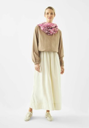 Vrouw met beige blouse, lange witte rok, roze sjaal met patroon en witte sneakers, staand tegen een witte achtergrond.