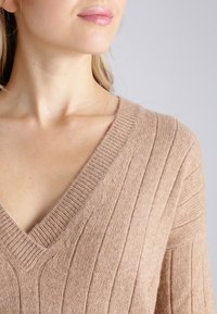 Gros plan sur une femme portant un pull côtelé beige à col en V, montrant le cou, la clavicule et une partie du visage avec des lèvres rose clair.