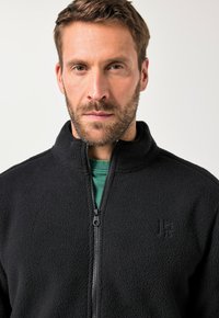 Chaqueta de forro polar negra con cuello alto y cierre de cremallera, que presenta una textura en la tela y un pequeño logo en el pecho izquierdo. Debajo, se ve una camisa verde.