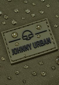 Tessuto verde oliva con una trama texturizzata, che presenta un'etichetta in gomma "JOHNNY URBAN" e gocce d'acqua chiare sulla superficie.