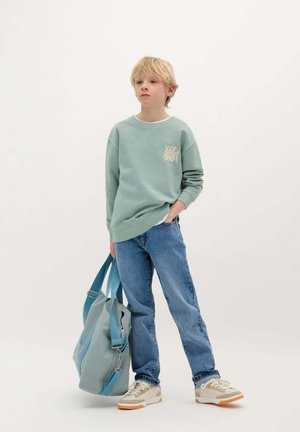 Felpa verde menta con logo sul petto, jeans denim blu e scarpe beige e grigie. Porta una borsa da viaggio azzurro chiaro.