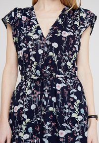 Blommönstrad klänning med marinblå bakgrund, med korta ärmar, V-ringning och elastisk midja. Lättviktsmaterial med mjuk textur.