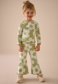 Grünes geripptes zwei-teiliges Outfit mit gro�ßen weißen Blumenmustern, bestehend aus weit geschnittenen Hosen und Volantsaumen, kombiniert mit cremefarbenen Sneakers.