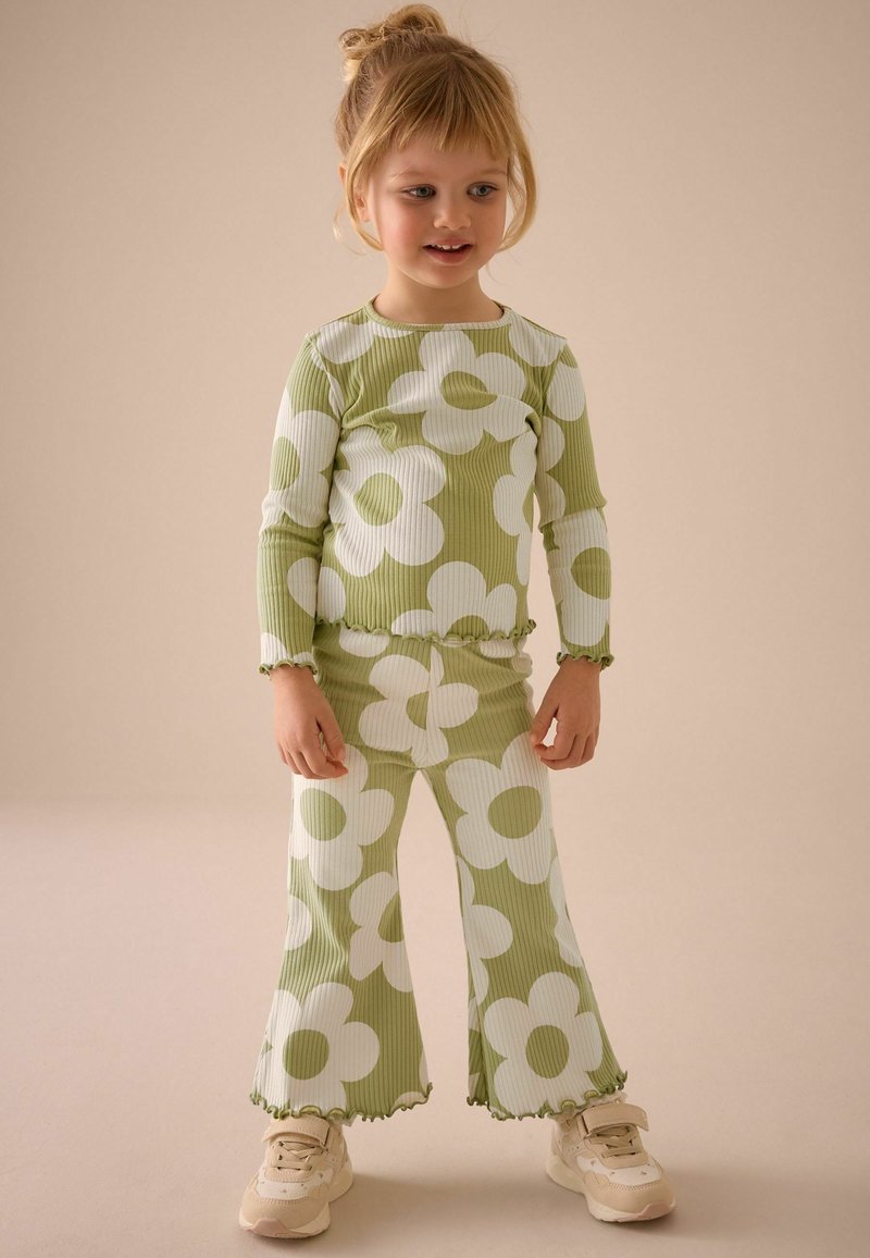 Grünes geripptes zwei-teiliges Outfit mit großen weißen Blumenmustern, bestehend aus weit geschnittenen Hosen und Volantsaumen, kombiniert mit cremefarbenen Sneakers.