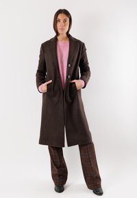 Cappotto di lana marrone con ampie rever, bottoni e tasche laterali, indossato sopra un maglione lavorato a maglia rosa e pantaloni a quadri marroni. Scarpe nere.