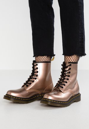 Metaliczne, różowe botki na sznurówki w kolorze różowego złota z czarnymi podeszwami i żółtym przeszyciem, noszone z czarnymi skarpetkami typu fishnet i krótkimi czarnymi jeansami.