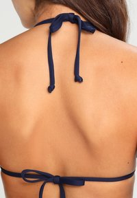 Marineblå bikini-top med tynne stropper knyttet bak. Glatt tekstur; minimalistisk design; avslører hud på skuldrene og øvre rygg.