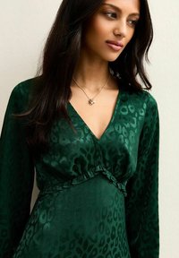 Vestido verde con estampado de leopardo con escote en V, mangas largas y detalles de volantes en la cintura. La modelo lleva un collar plateado sencillo. Textura suave.