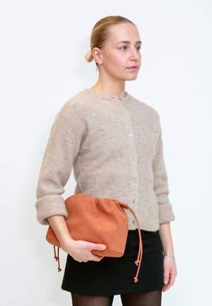 Kvinde med blondt hår i en beige cardigan og sort nederdel holder en stor orange taske mod en ensfarvet hvid baggrund.