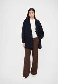 Palton supradimensionat bleumarin cu reveruri mari, asortat cu un top alb cu model nervurat și pantaloni maro cu picioare largi. Pantofii sunt cu toc ascuțit, albi.