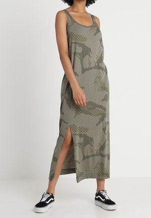 Persona con vestido midi sin mangas con estampado de camuflaje y abertura lateral, combinado con zapatillas negras y blancas sobre fondo claro.