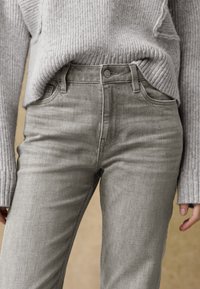 Graue Jeans mit taillierter Passform, ausgestattet mit einem Knopfverschluss und Fronttaschen. Getragen mit einem hellgrauen Rippensweater.