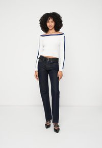 Fehér, bordázott, vállnélküli pulóver kék csíkkal, sötétkék farmernadrággal és fekete, hegyes orrú magassarkú cipővel, bokapánttal párosítva.