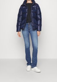 Mörkblå puffjacka med en glansig yta, dragkedjestängning och hög krage; i kombination med blå jeans och vita sneakers.