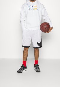 Felpa bianca con testo colorato "BROOKLYN, NEW YORK" e pantaloni corti neri con lo swoosh bianco della Nike, abbinati a scarpe nere e calzini rossi.