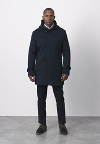 Schwarzer, kniehoher Mantel mit Kapuze, ausgestattet mit Reißverschluss, Seitentaschen und gerippten Bündchen. Wird über dunklen Hosen und Stiefeln getragen.