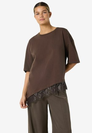 Femme portant un haut marron ample à manches courtes avec un ourlet en dentelle asymétrique et un pantalon marron large assorti, posant sur un fond blanc.
