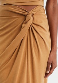 Hnědá, elastická sukně s uzlovým detailem v pase a splývavou texturou, vytvářející jemné záhyby. Detailní záběr na design.
