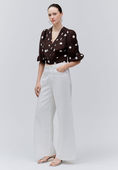 Femme debout, main dans la poche, vêtue d'un chemisier noir à pois blancs, d'un pantalon large blanc, portant des boucles d'oreilles anneaux et des sandales argentées.