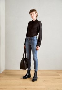 Svart langermet skjorte, blå skinny jeans, svarte ankelstøvler og en brun tote bag. Enkelt design med knappdetaljer og glatte teksturer.