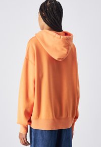 Sudadera naranja con un corte relajado, capucha grande y puños acanalados. Fabricada con una tela suave de textura lisa. Combinada con jeans de mezclilla azules.