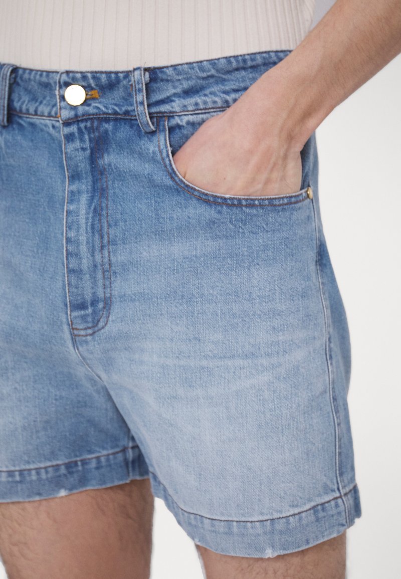 Shorts en denim de couleur bleu clair avec une coupe classique. Parmi les caractéristiques, on trouve une poche avant, des coutures visibles et un bouton en métal à la taille.