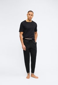 Άνδρας όρθιος, ξυπόλητος, φορώντας μαύρο πουκάμισο με κοντά μανίκια και μαύρο παντελόνι jogger με ριγέ μανσέτες, σε λευκό φόντο.