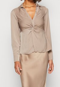 Femme portant un blouse en satin beige avec un détail avant tordu, à manches longues, assortie à une jupe en satin et un collier en chaîne dorée.