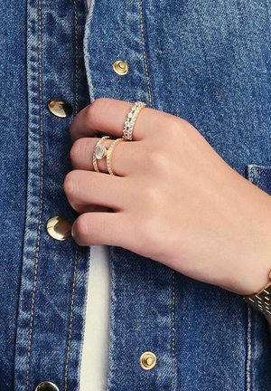 Tre ringar på en hand: en guld med en klar sten, en guld med små diamanter, en tunn guldring. Handen vilar på en denimjacka med guldfärgad knappar.
