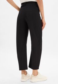Black high-waisted trousers with a relaxed fit, featuring two back pockets and a smooth, stretchy fabric. Worn with off-white sneakers.