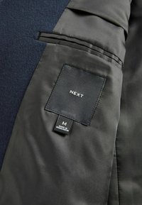 Interior de abrigo azul marino oscuro con forro negro suave, presentando una etiqueta marcada "NEXT" y una etiqueta de tamaño "M". Costuras detalladas visibles.